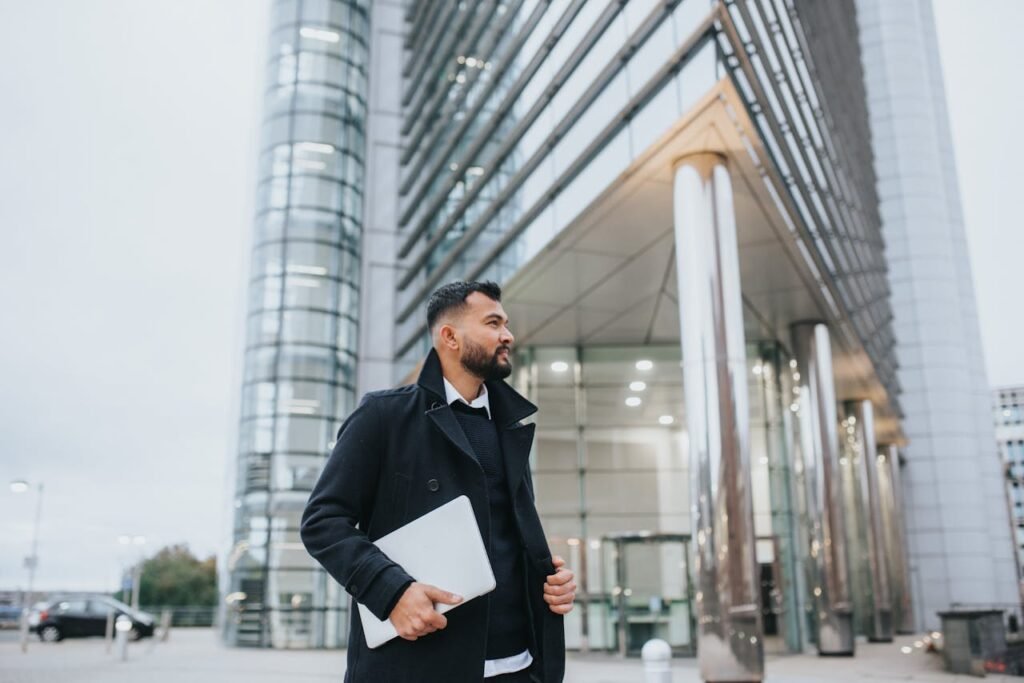 Dreamy unshaven ethnic male executive in stylish wear with netbook looking away against contemporary construction in city